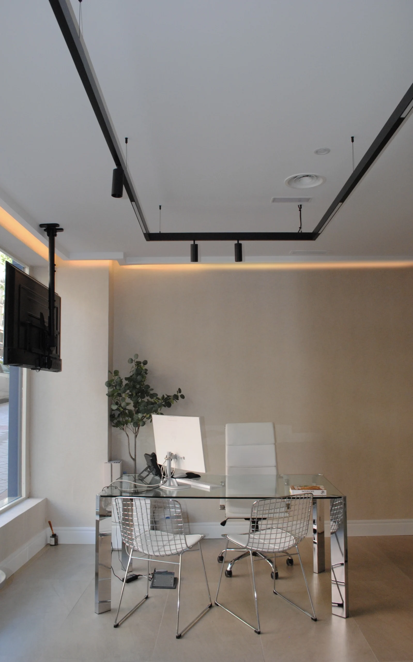oficina minimalista con mesa de cristal, silla ejecutiva blanca, dos sillas metálicas, iluminación lineal negra y luz indirecta cálida.