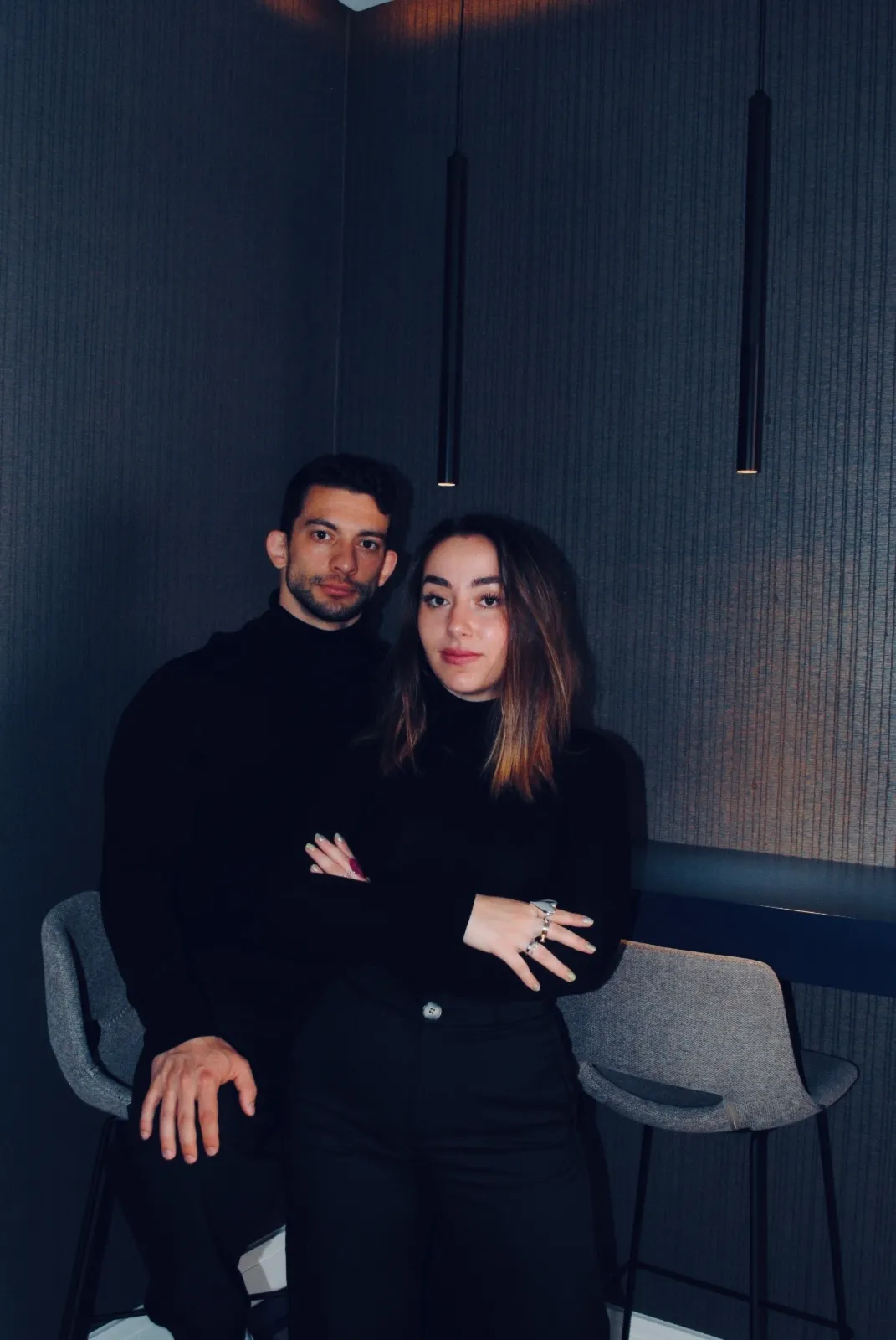 4f5e2d03-f9d3-4349-b21e-e686d1f9e317 Borja Yaques y Helena Alarcón, los dos cofundadores de BYE Interior posan en su estudio, vestidos de negro, junto a una barra alta con taburetes grises y luminarias colgantes minimalistas